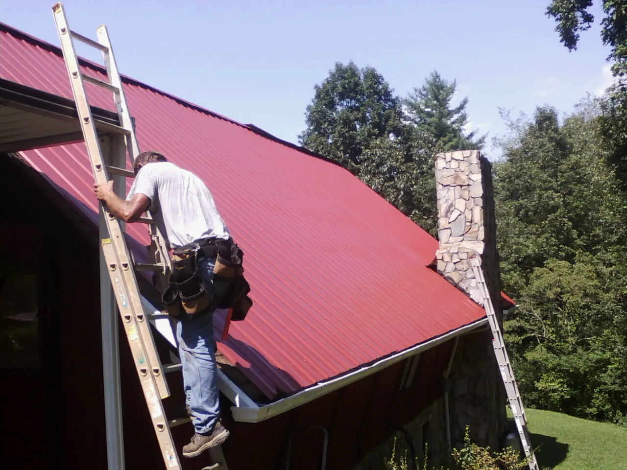 Roofer on a ladder beside a red metal roof and stone chimney — Soffit & Fascia Repair by Roofing Brothers in Illinois City